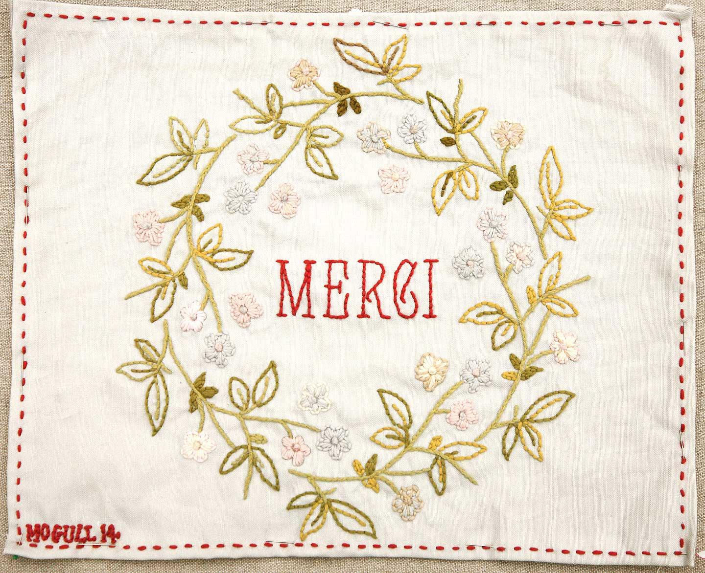 Embroidered fabric with floral wreath and 'MERCI' text on a off-white background