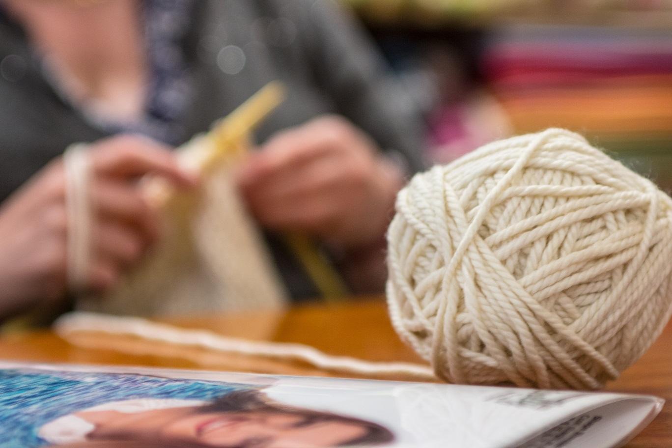 Ball of yarn and knitting needles on a table with a blurred person in the background.