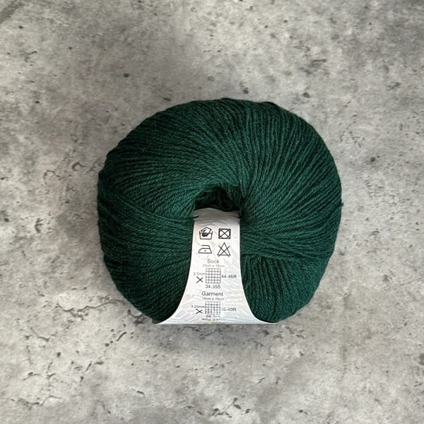 A ball of dark forest green yarn with a white care label, placed on a textured gray concrete surface.