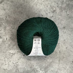 A ball of dark forest green yarn with a white care label, placed on a textured gray concrete surface.