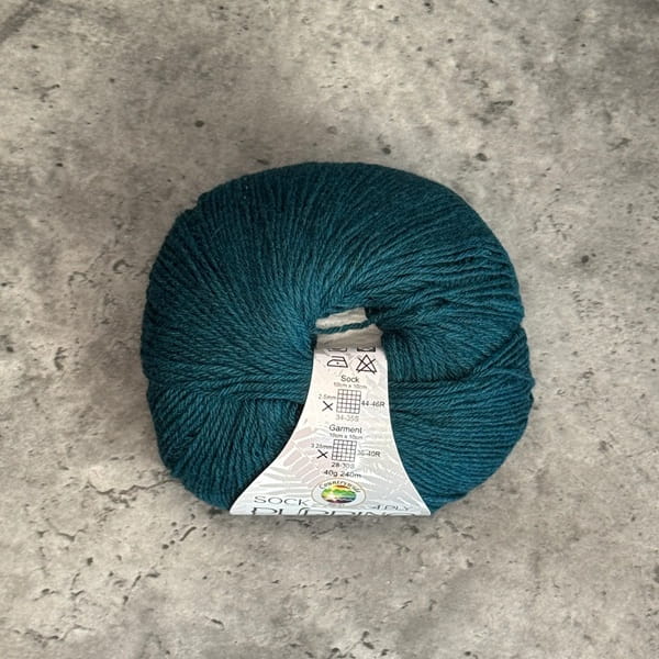 A ball of deep teal yarn with a white paper label, placed on a textured gray concrete surface.