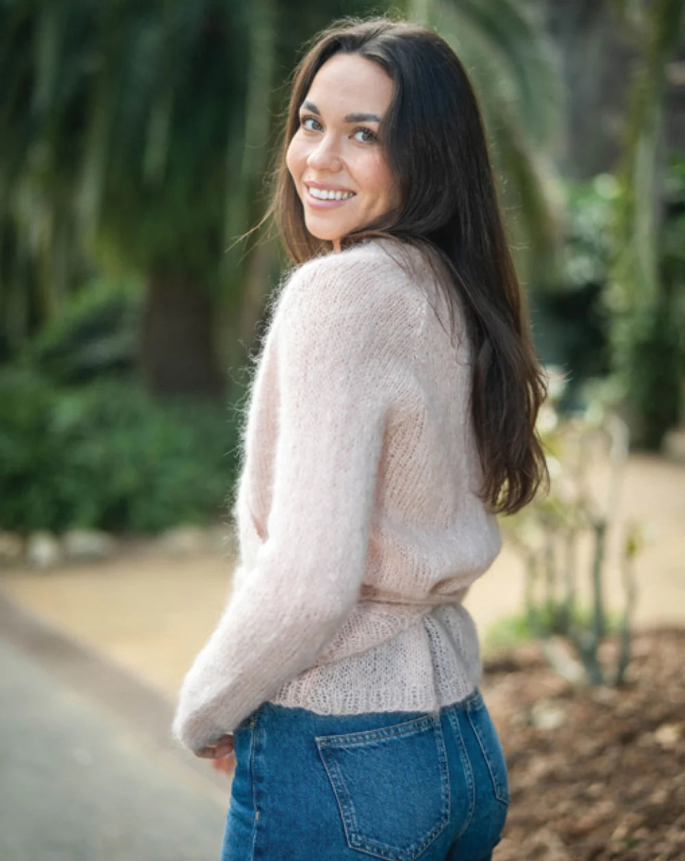 Woman wearing a light pink sweater and blue jeans standing outdoors with greenery in the background