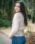 Woman wearing a light pink sweater and blue jeans standing outdoors with greenery in the background