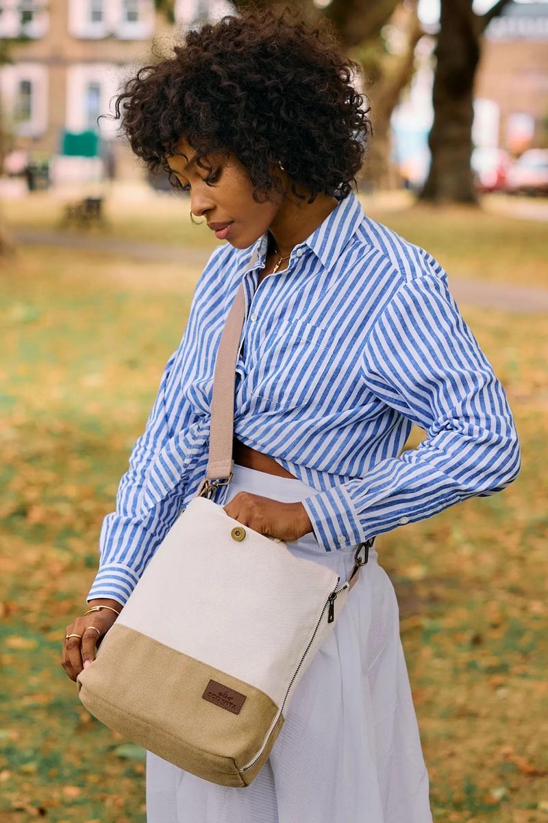 Woman in a blue striped shirt and white skirt holding a beige and brown bag in a park.
