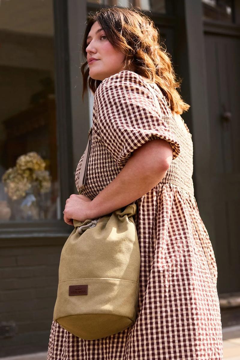 Woman in a checkered dress holding a beige bucket bag outdoors.