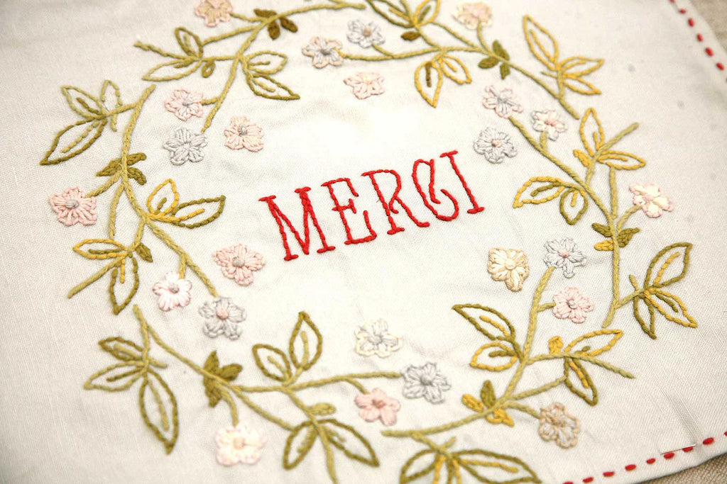Embroidered text 'MERCI' surrounded by floral patterns on a off- white fabric background.