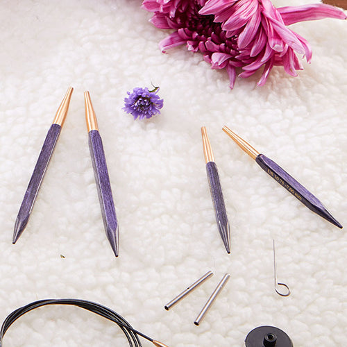 Knitpro purple knitting needle tips with gold accents on a textured white surface with pink flowers.