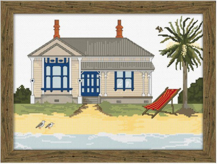 Cross-stitched design of a beach house with a palm tree and lounge chair, framed in wood.