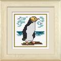 Cross-stitch kit depiction of a yellow-eyed penguin on white Aida fabric with a blue ocean and sky in the background, framed in a beige frame.