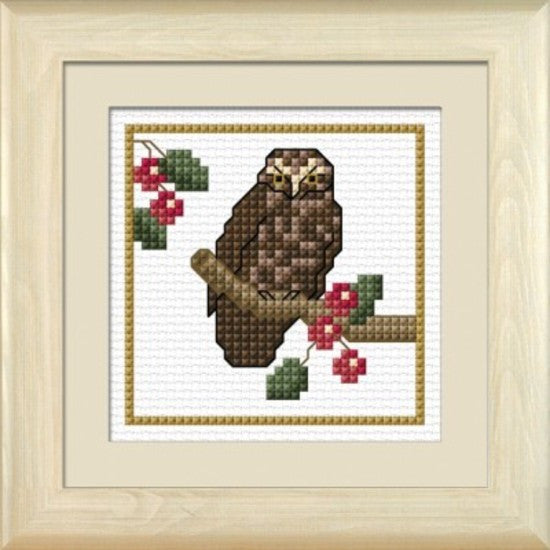 Cross-stitch kit depicting a Morepork owl perched on a branch with red berries, framed in a light-colored frame.