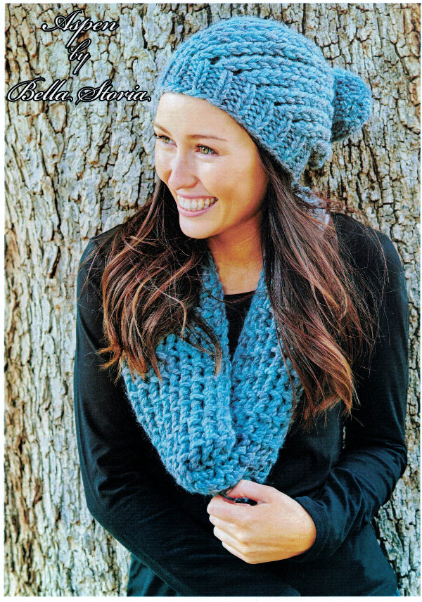 A woman wearing a blue chunky knit hat and a black cowl, standing outdoors against a tree background.