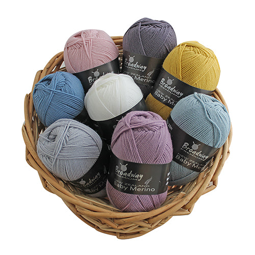 A basket containing multiple skeins of New Zealand merino yarn in various shades of blue, gray, pink, silver, and white, labeled as 'Broadway Baby Merino'.